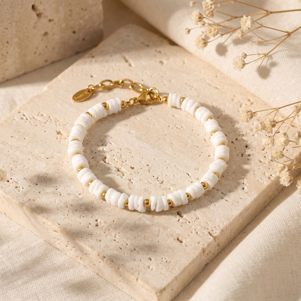 White disc-bead bracelet with gold-tone spacers arranged on a beige stone slab beside dried flowers.