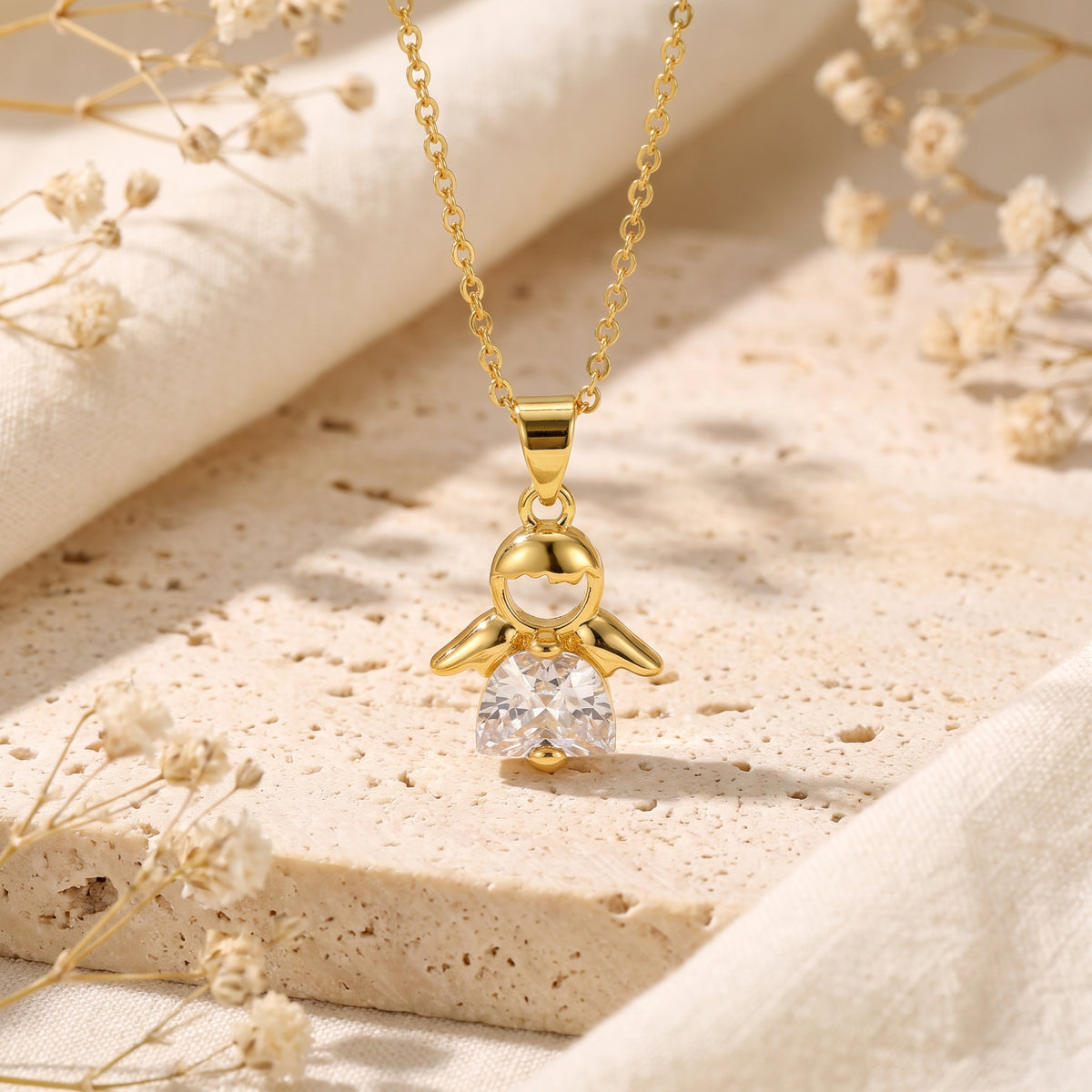 Front-facing gold-tone angel pendant with a clear cubic zirconia gown, suspended over a textured stone surface.