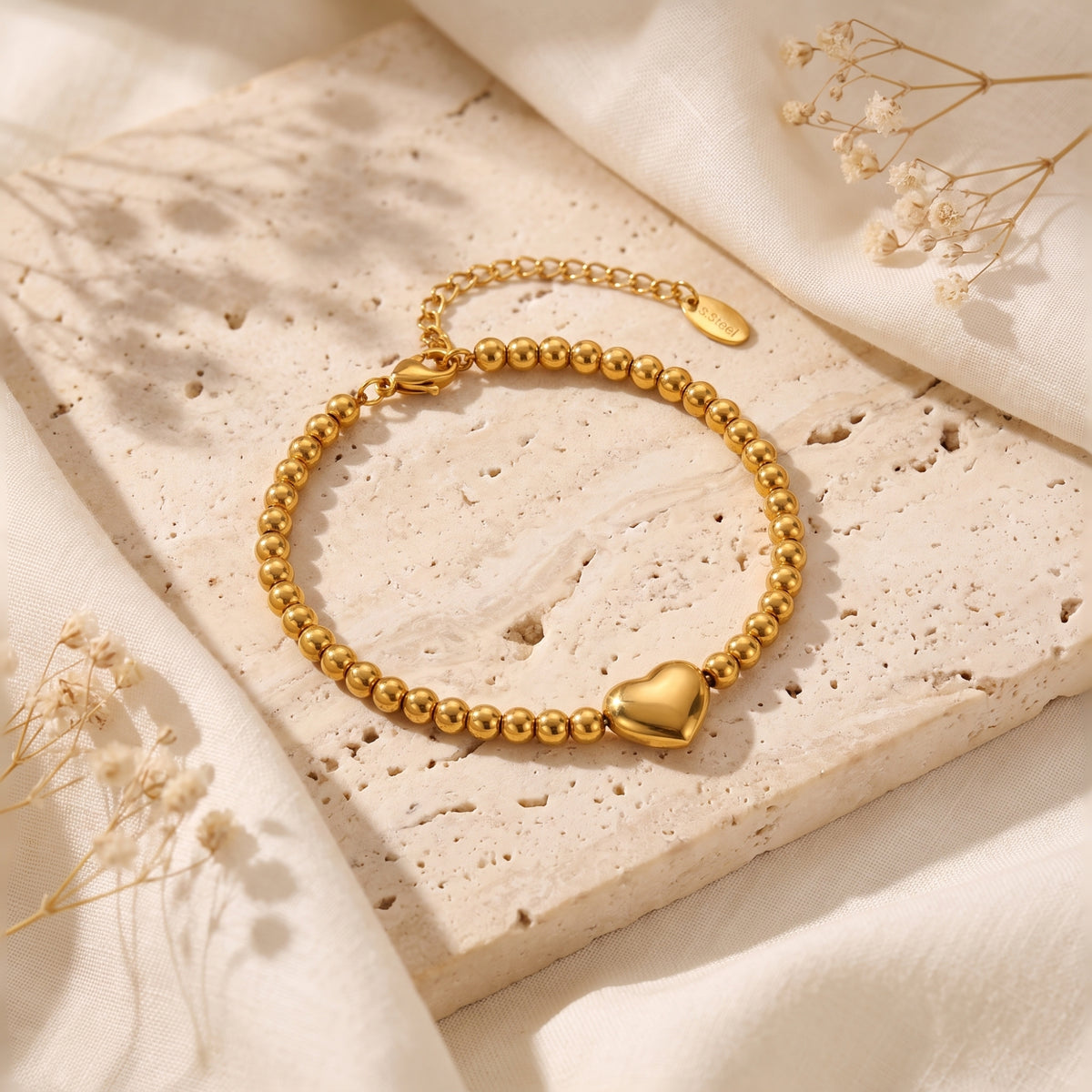 Gold-tone beaded bracelet with a smooth puffed heart charm, displayed on a textured stone surface with cream fabric.