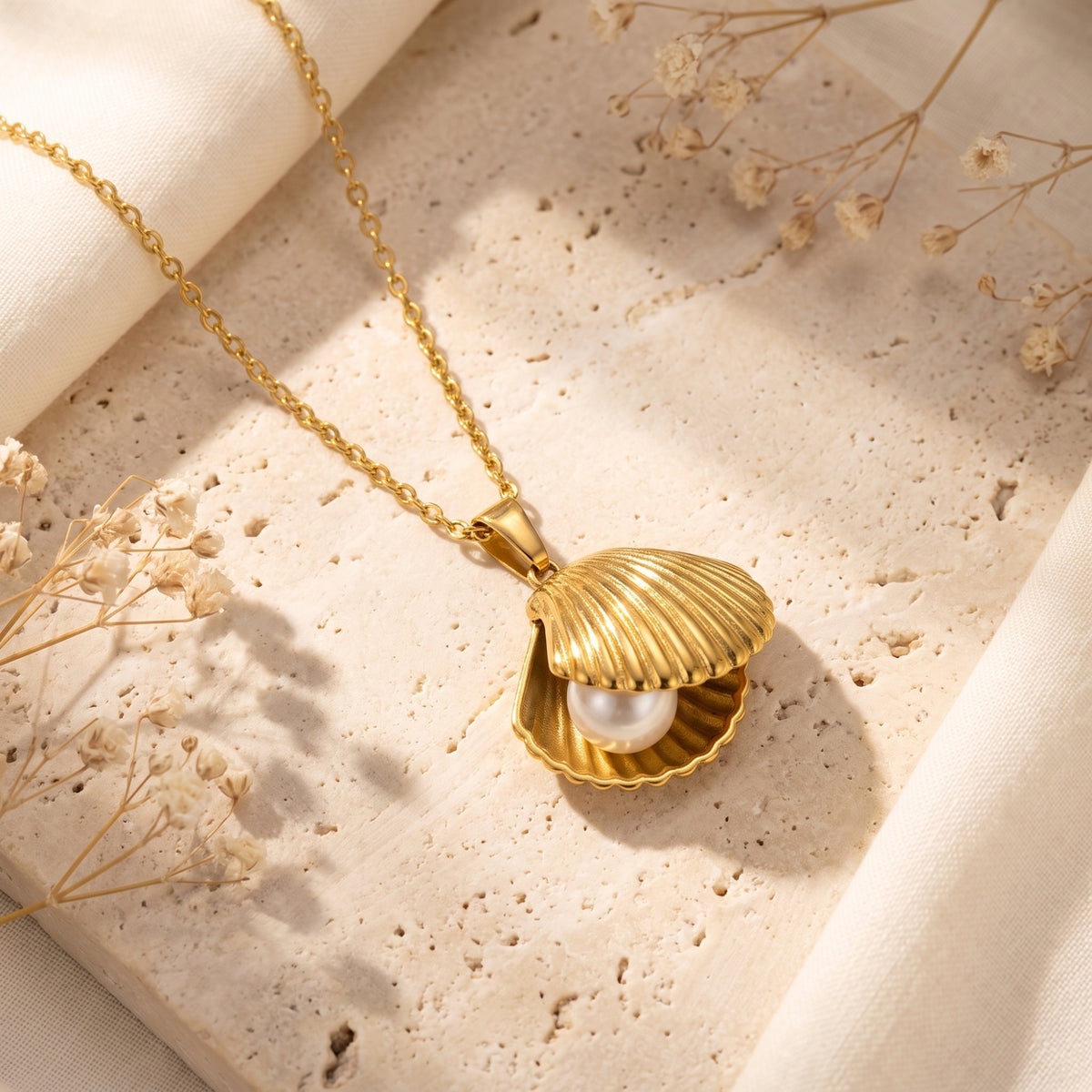 Gold-tone open shell pendant with a white pearl, chain, and clasp arranged on a pale textured stone surface.