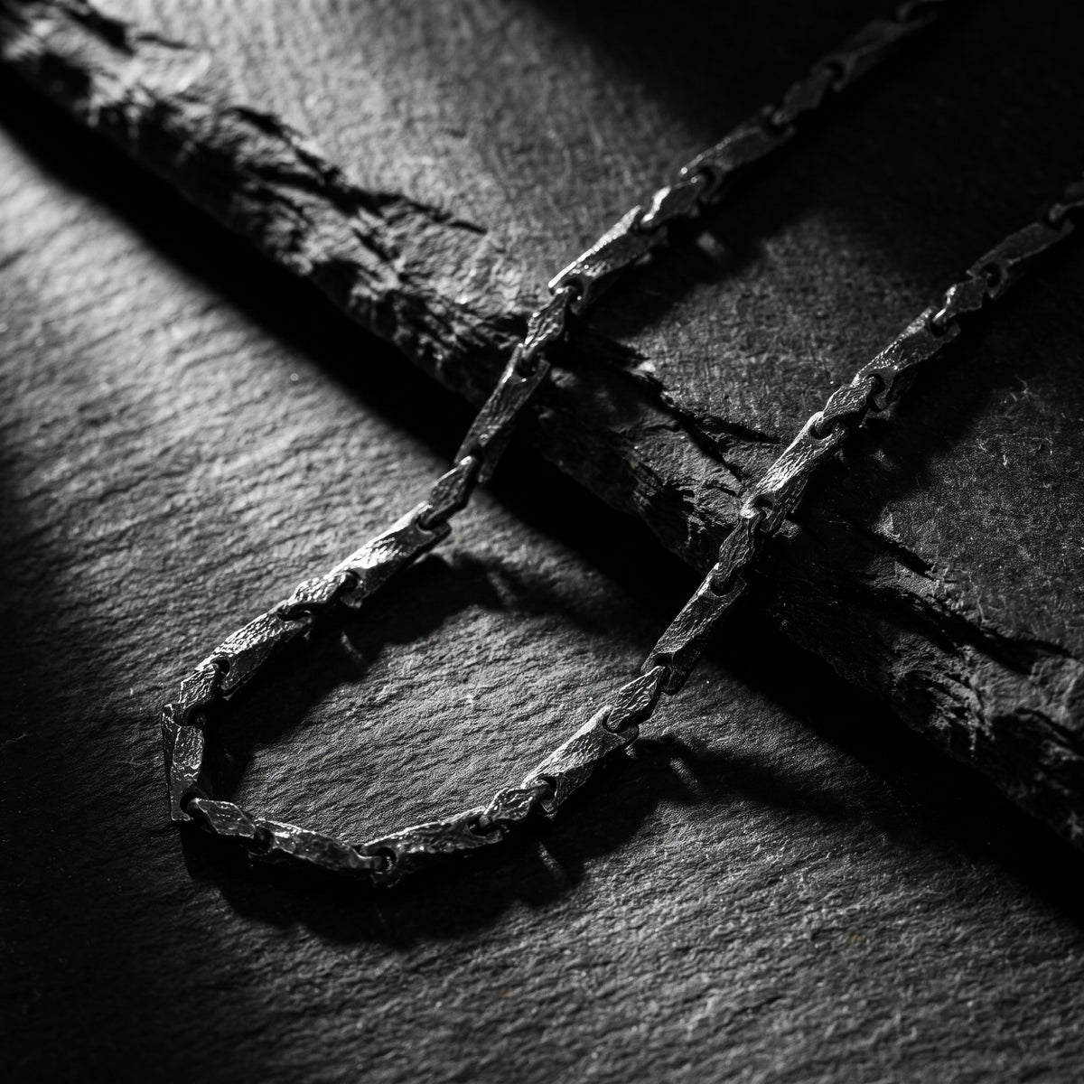 Textured silver bar link chain resting on a dark slate surface in dramatic lighting.