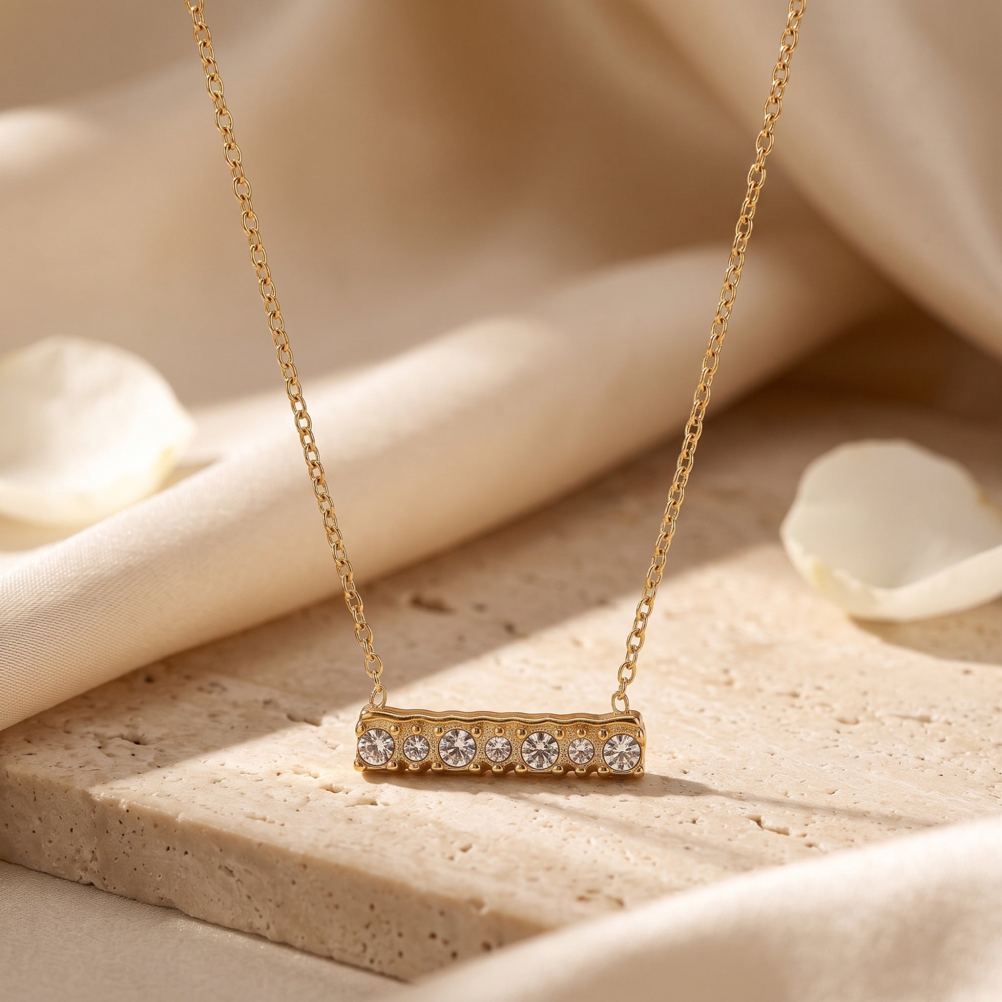 Gold bar pendant necklace with seven round cubic zirconia stones on a textured stone and fabric background.