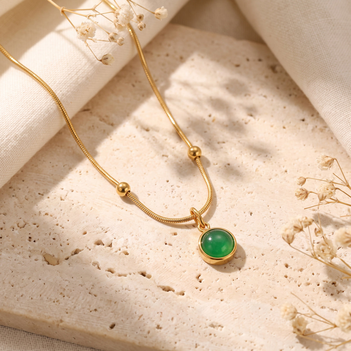 Gold-tone snake chain necklace with sphere stations and bezel-set green cabochon pendant on a stone slab.
