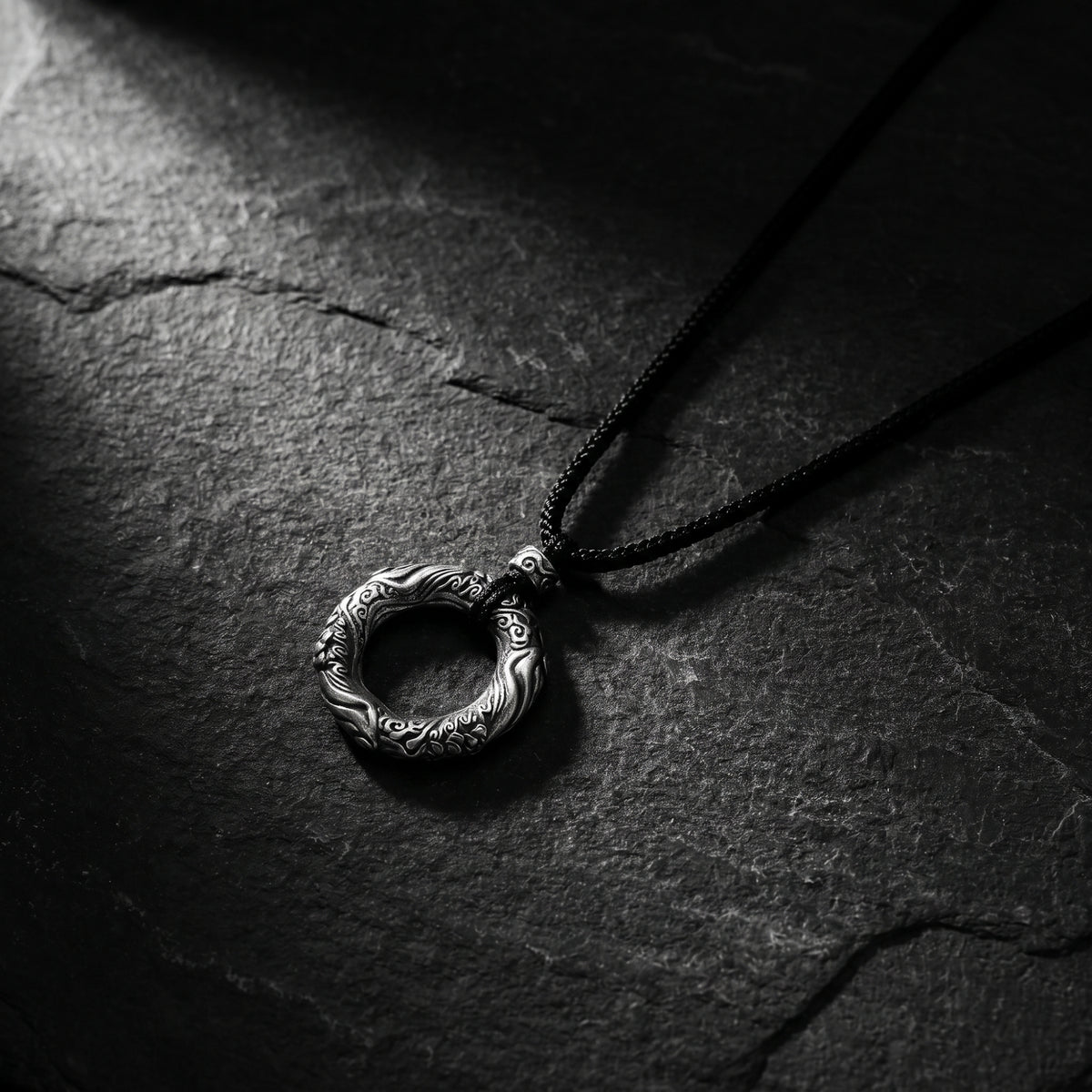 Carved circular silver ring pendant attached to a black cord, lying on dark slate stone.