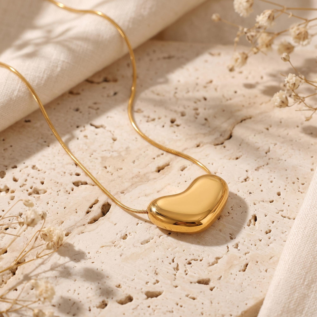 Close-up of a gold-tone puffy heart pendant on a snake chain, set on textured stone with dried flowers.