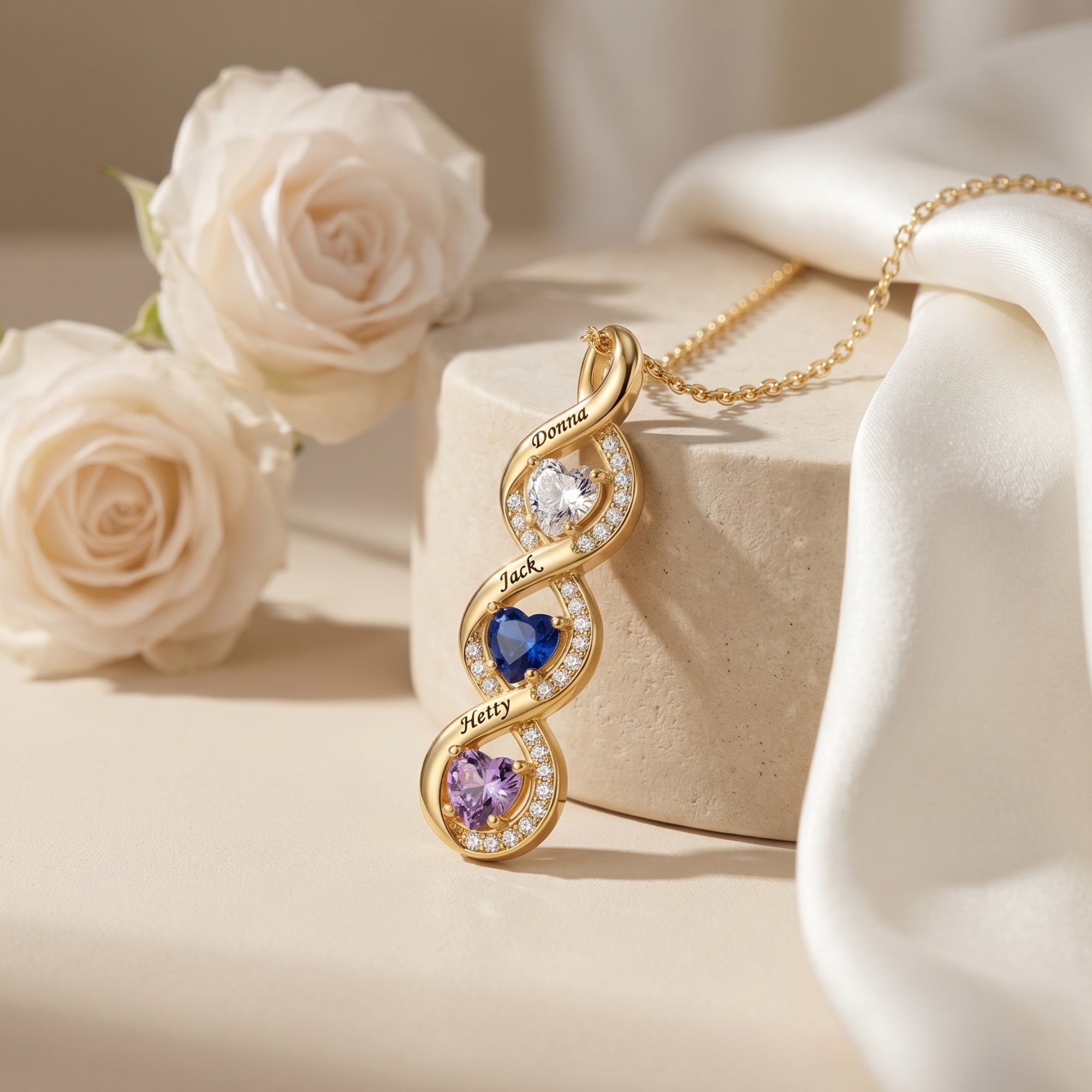 Gold infinity necklace with heart birthstones and names on a stone pedestal with roses.