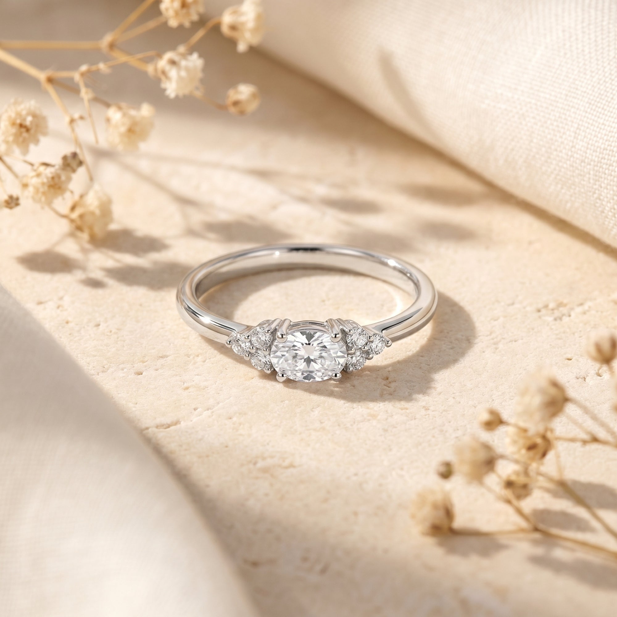 Oval cut moissanite engagement ring in sterling silver on a stone surface with dried flowers.