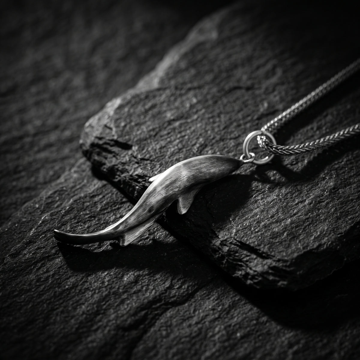 Silver shark pendant lying horizontally on dark slate, with its braided chain attached