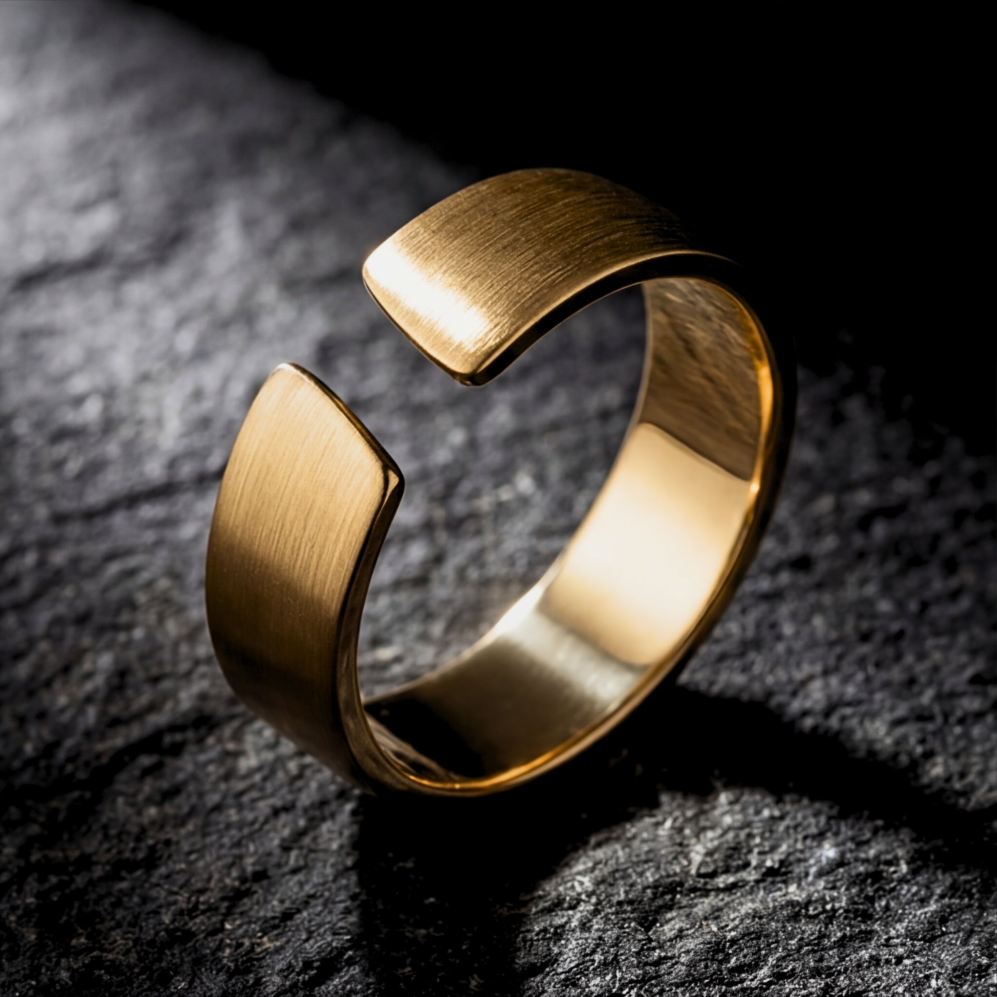 Three-quarter view of a gold stainless steel gap ring with a brushed finish on a dark stone background.
