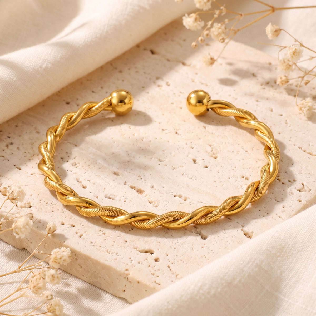 Gold-tone twisted cuff bracelet with smooth ball ends, displayed flat on a beige stone surface.