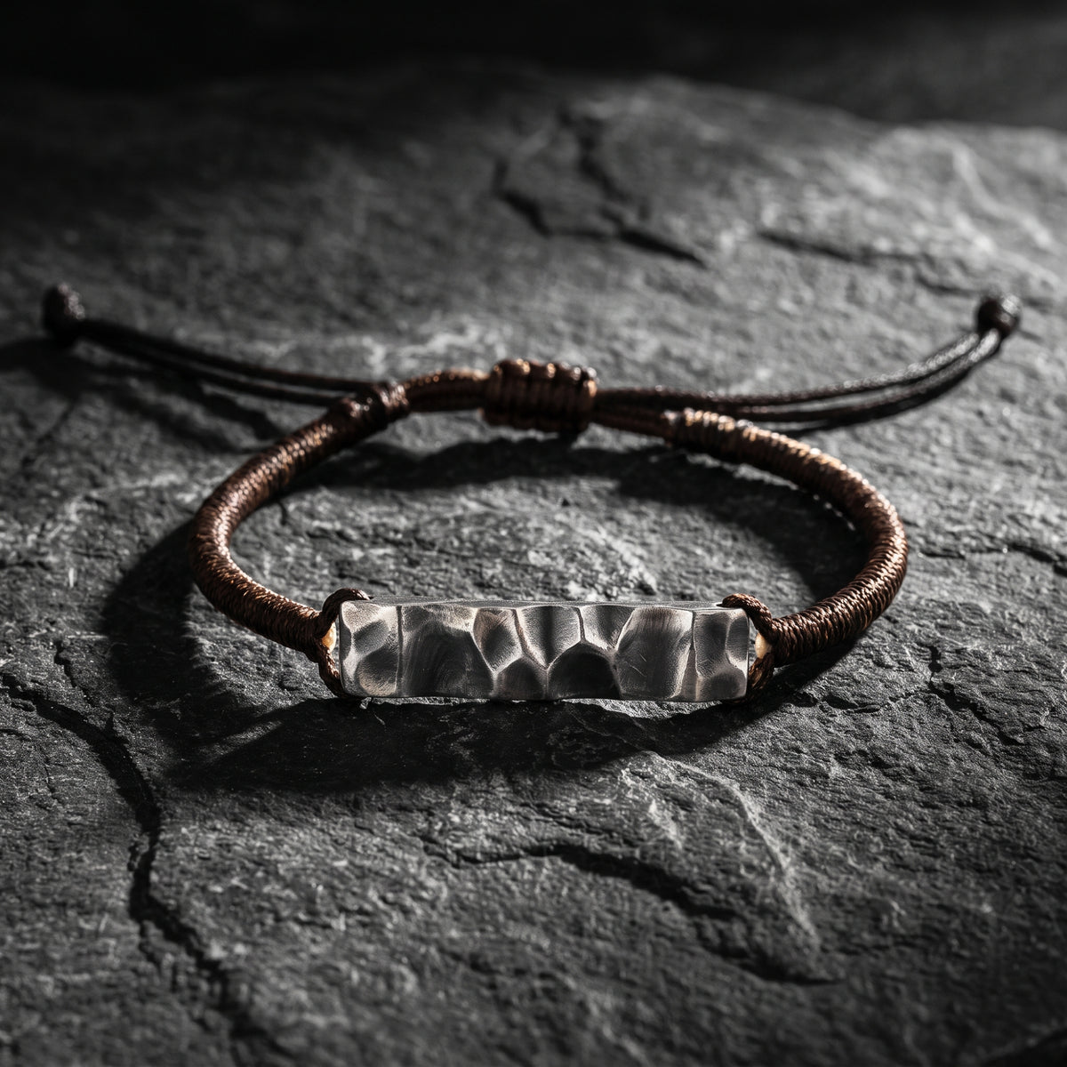 Hammered bar silver bracelet with brown woven adjustable cord, resting flat on dark slate stone.