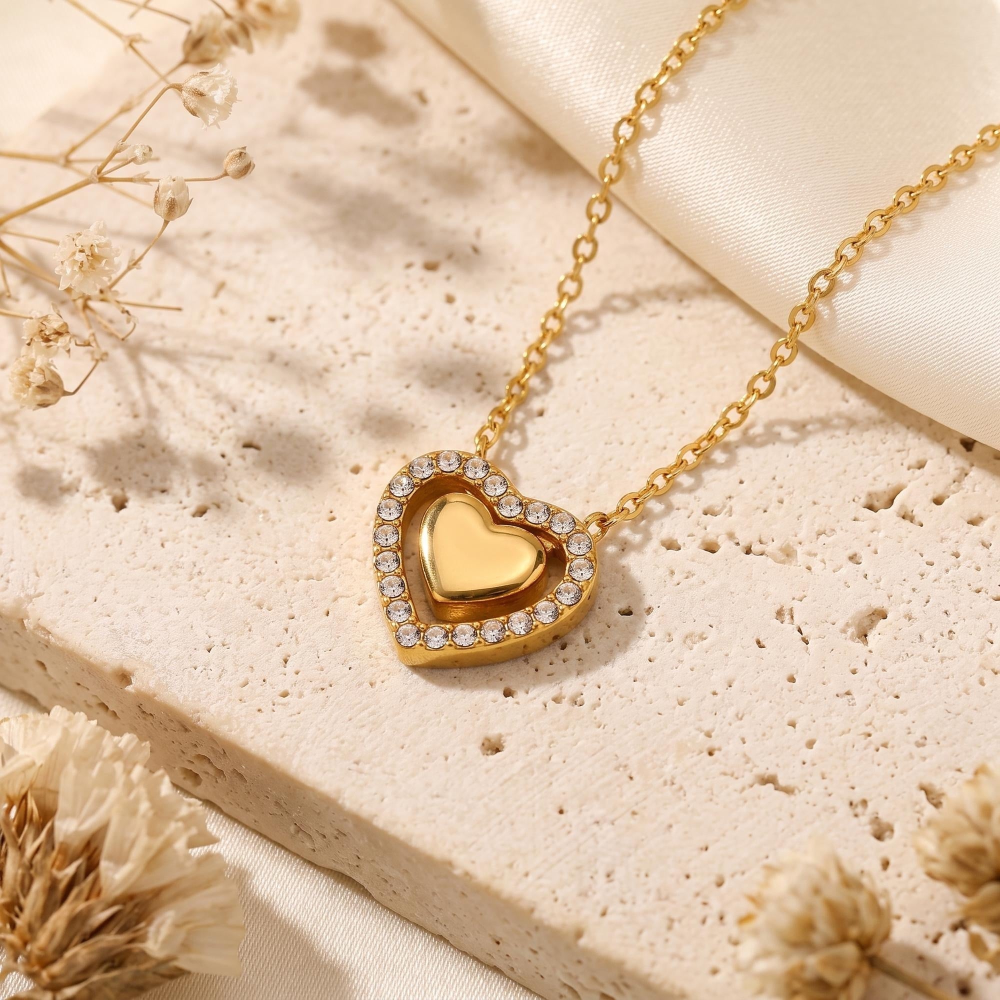 Gold double heart pendant necklace with cubic zirconia accents on a textured stone lifestyle background.