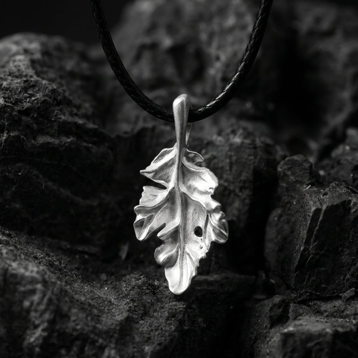 Front close-up of a silver oak leaf pendant on a black cord, suspended over dark rocks.