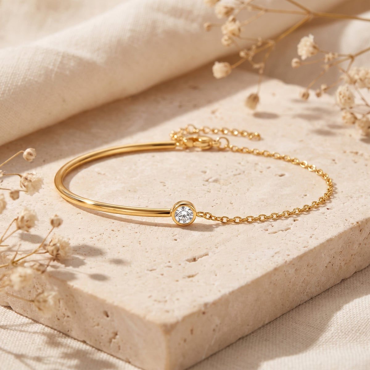 Gold-tone bezel station cuff bracelet with a round clear stone, displayed on a beige textured stone block with dried flowers.