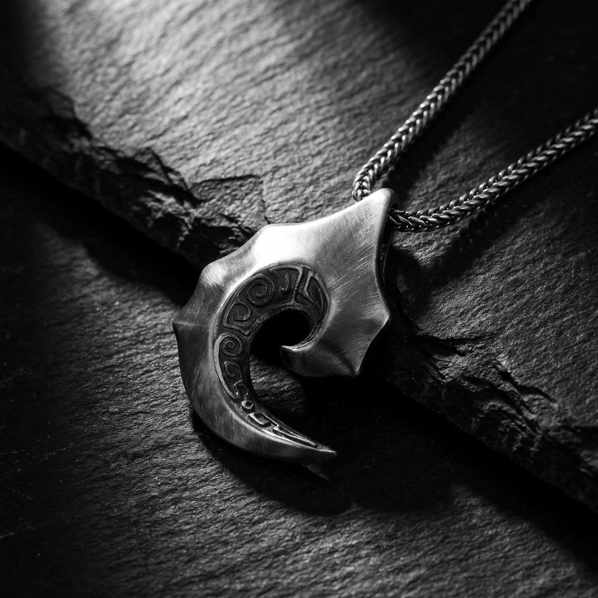 Close-up of a silver wave scroll pendant with carved detailing resting on dark slate.