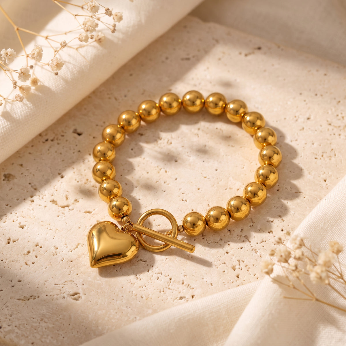 Gold-colored beaded bracelet with a puffy heart charm and toggle clasp resting on a stone surface.