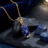 Faceted pear natural lapis lazuli pendant necklace in solid gold on a dark rock with raw lapis stones.