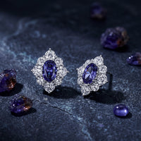 Oval natural tanzanite drop earrings in white gold standing on a dark stone surface with raw gemstones.