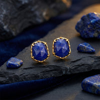 Gold hexagon stud earrings with natural blue lapis lazuli stones on a dark slate background with raw gemstones.