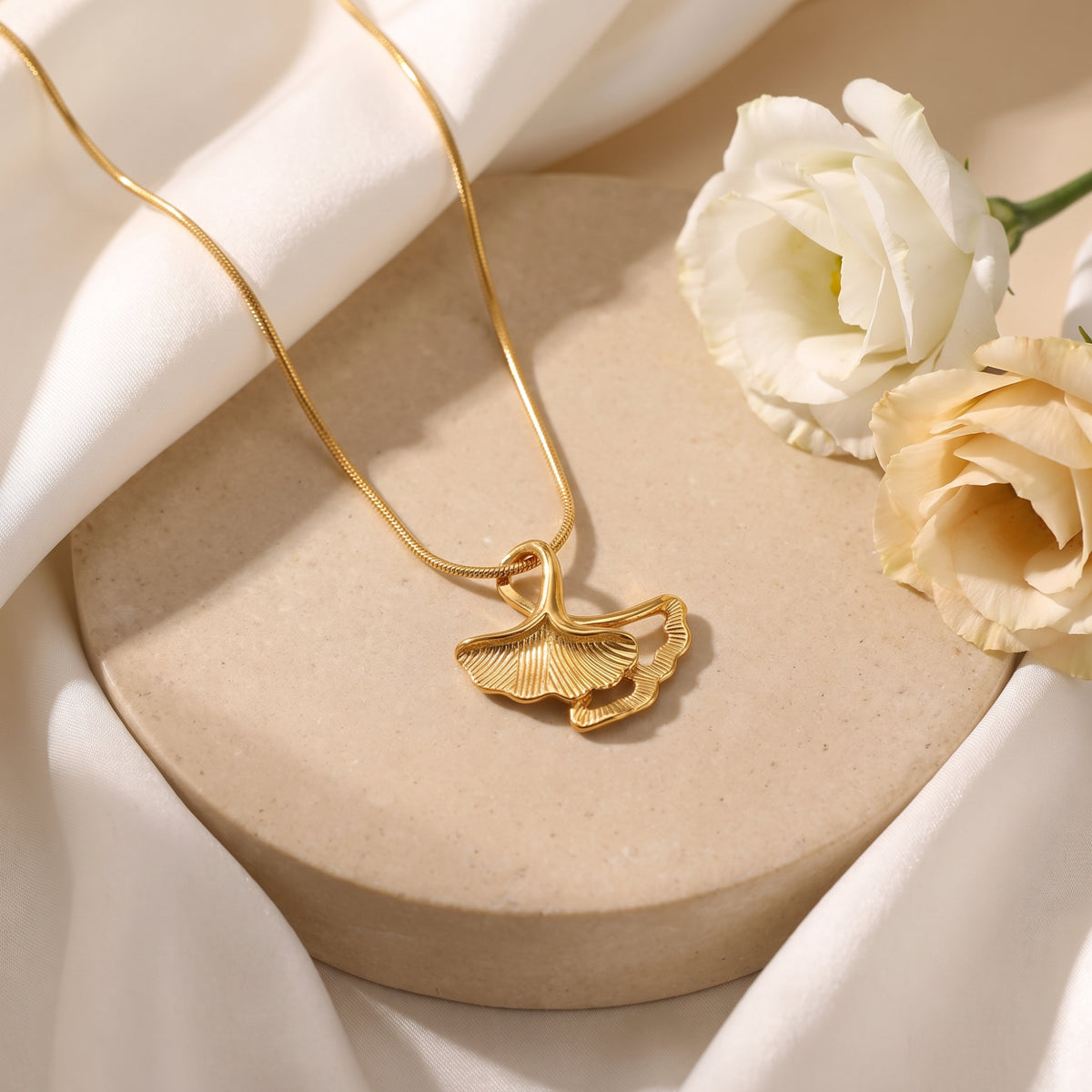 Gold-tone double ginkgo leaf pendant necklace displayed on a beige stone platform with white roses.