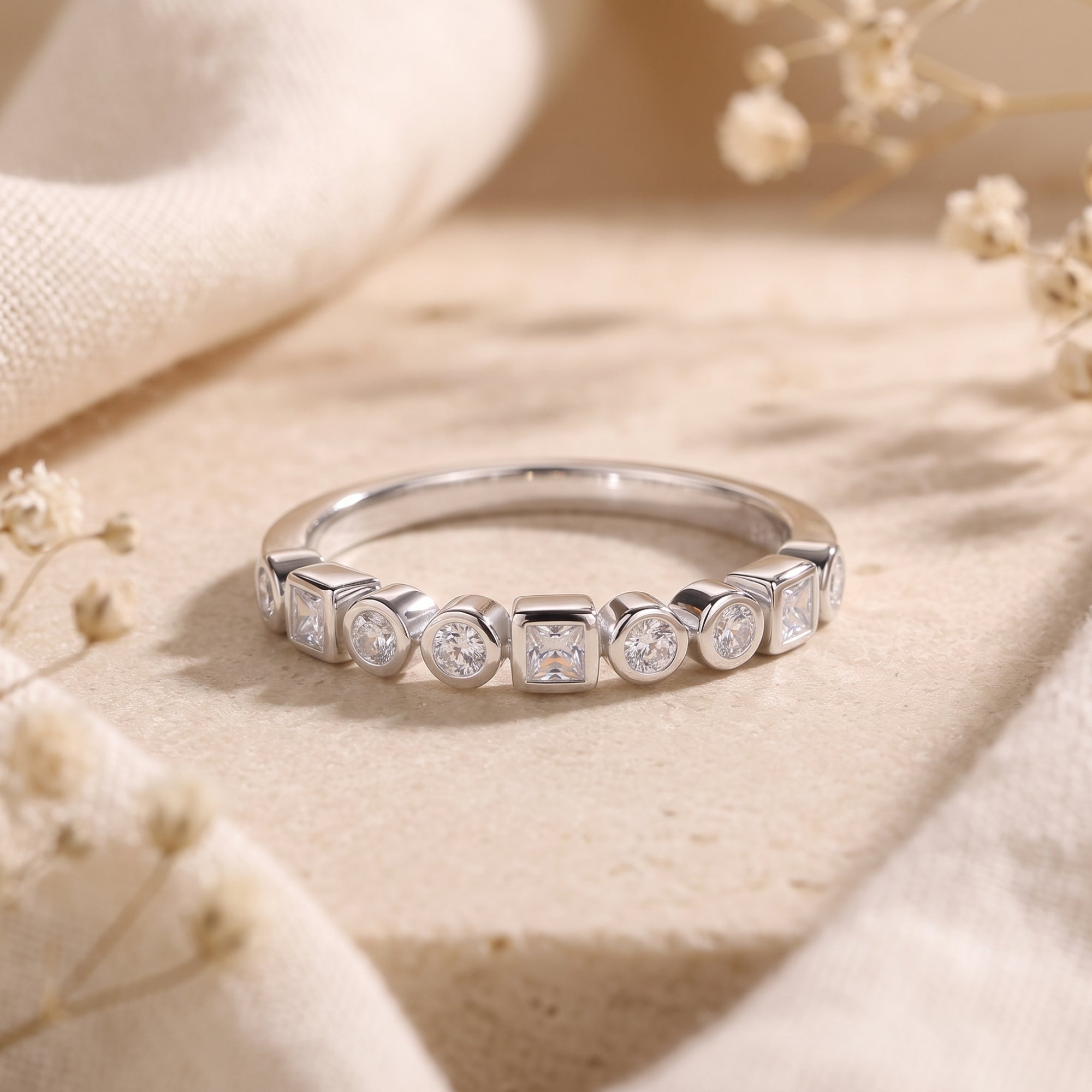 Silver eternity band with alternating round and princess-cut stones on a stone surface with flowers.