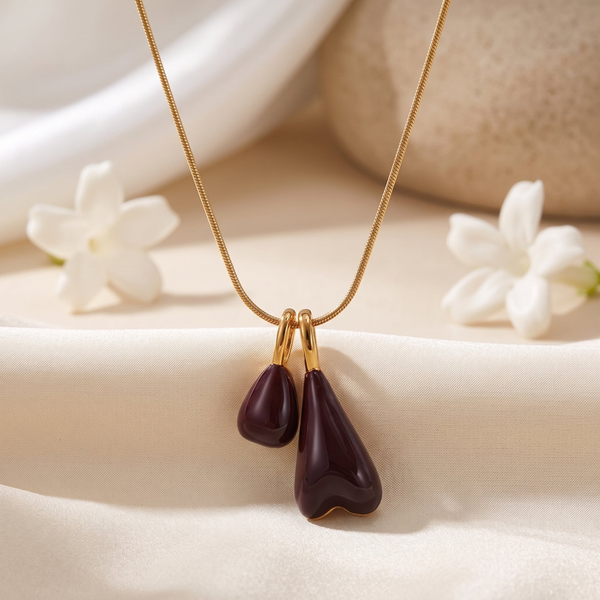 Marais gold stainless steel pendant necklace with burgundy double drops styled on white fabric with flowers.