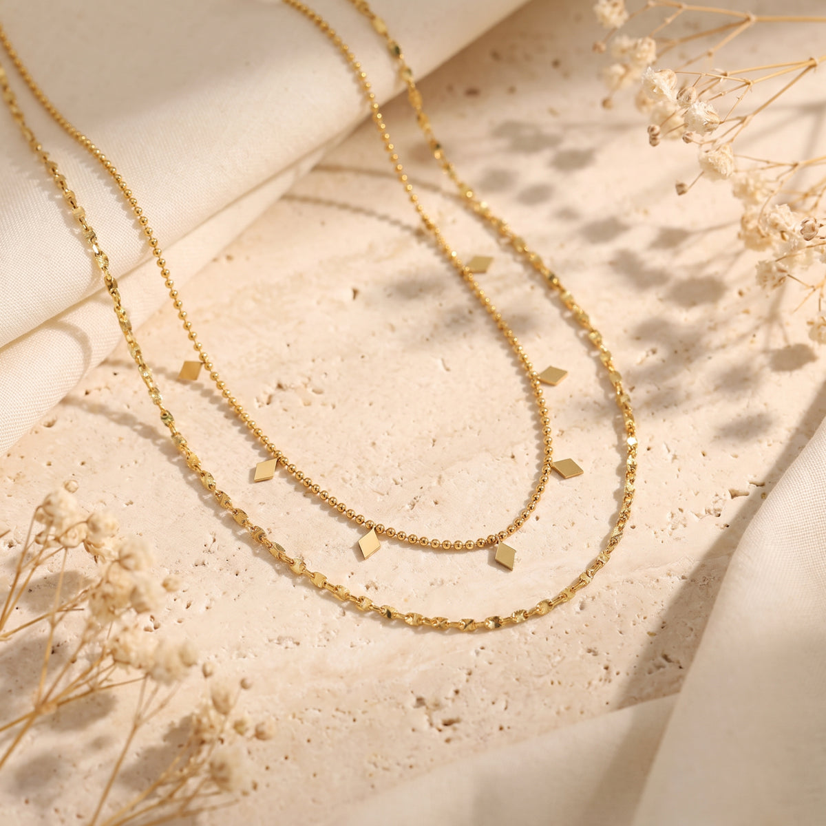 Gold-tone layered necklace with a beaded inner chain and diamond-shaped charms, arranged on stone and cream fabric.
