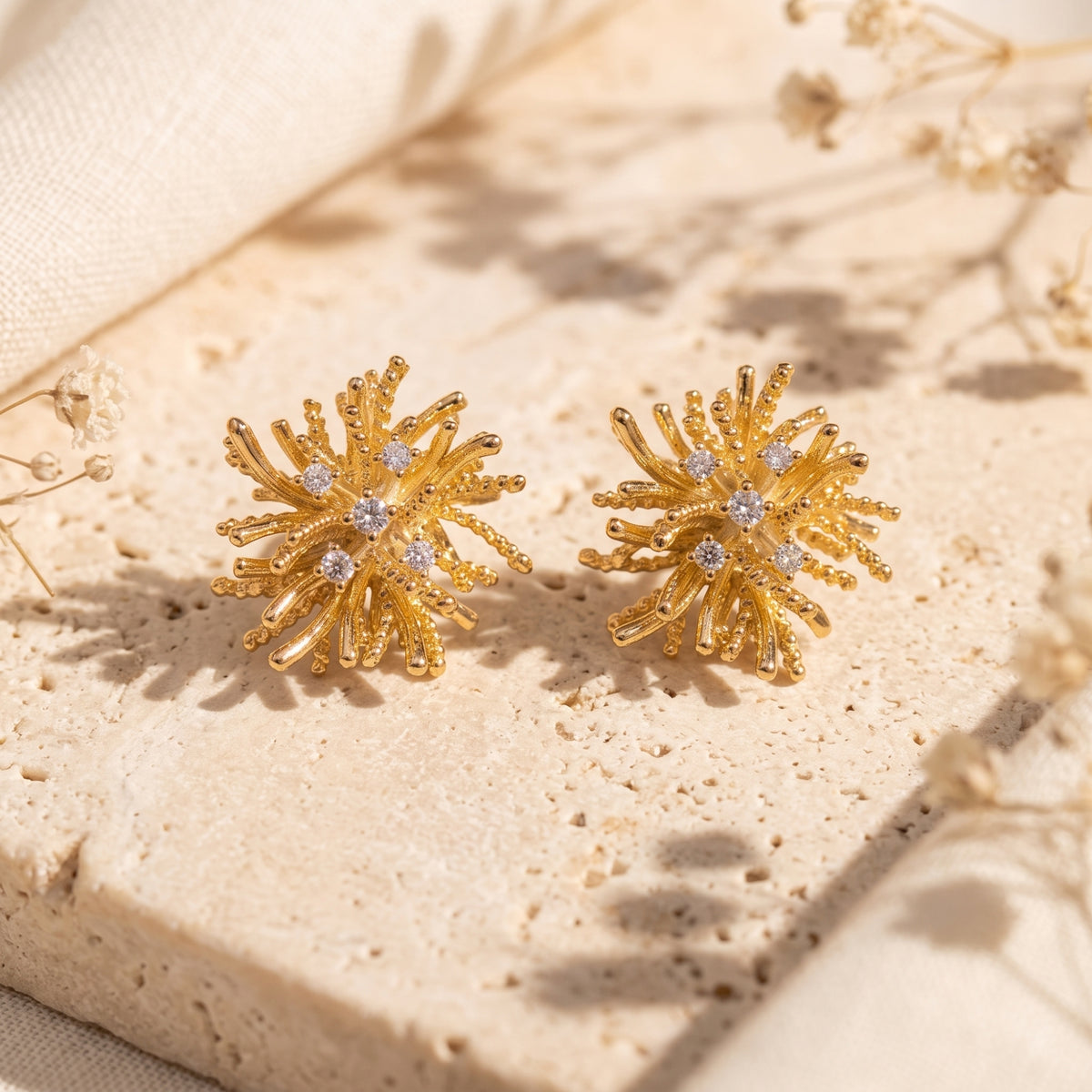 Gold-tone starburst stud earrings set with clear stones resting flat on a beige stone surface.