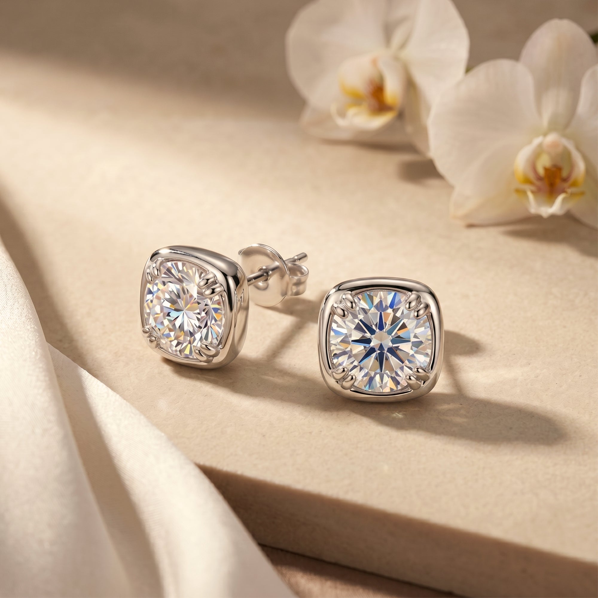 Silver Gabriella cushion-halo moissanite stud earrings on a stone surface with white orchids.