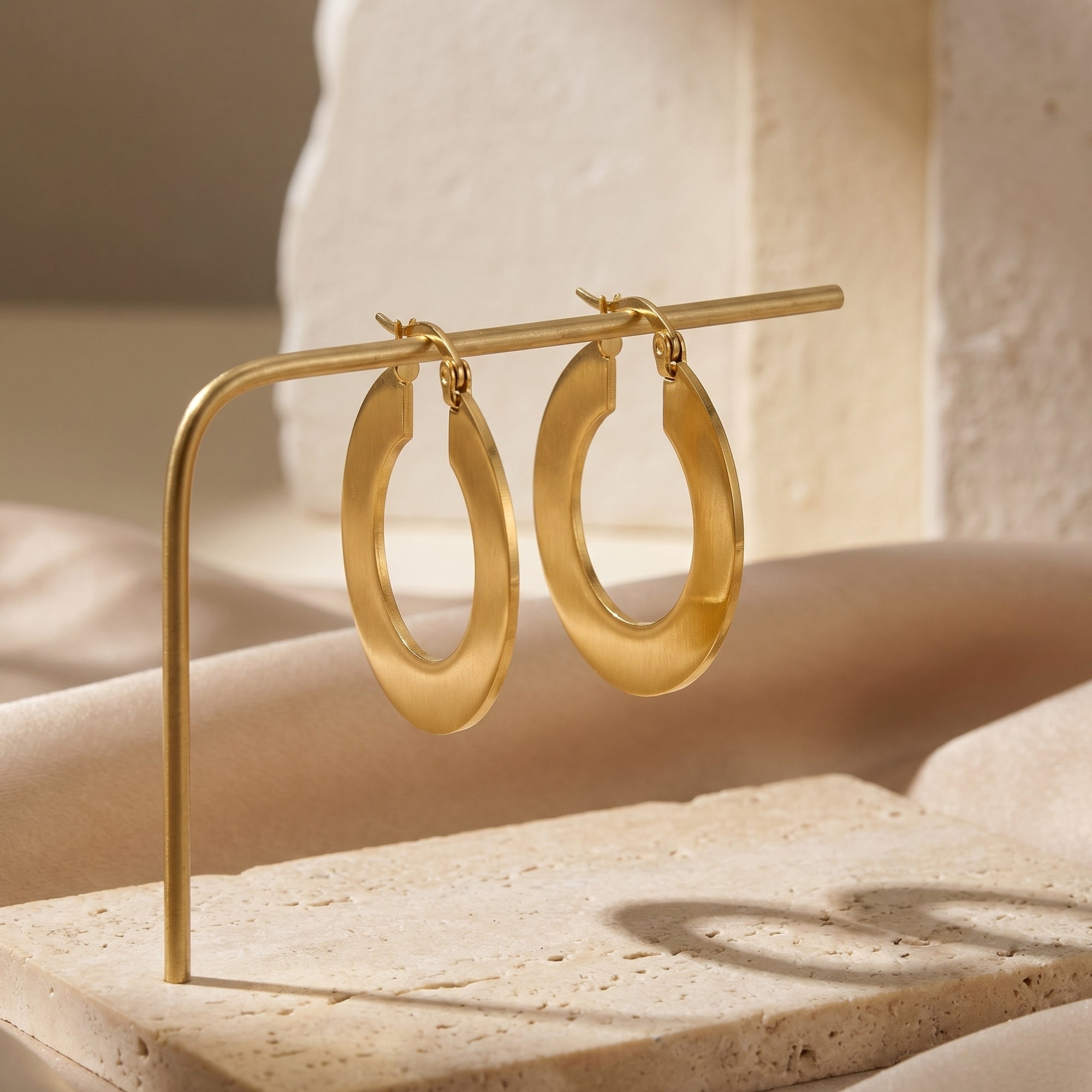 Matte 18K gold PVD stainless steel hoop earrings displayed on a gold stand with a stone background.