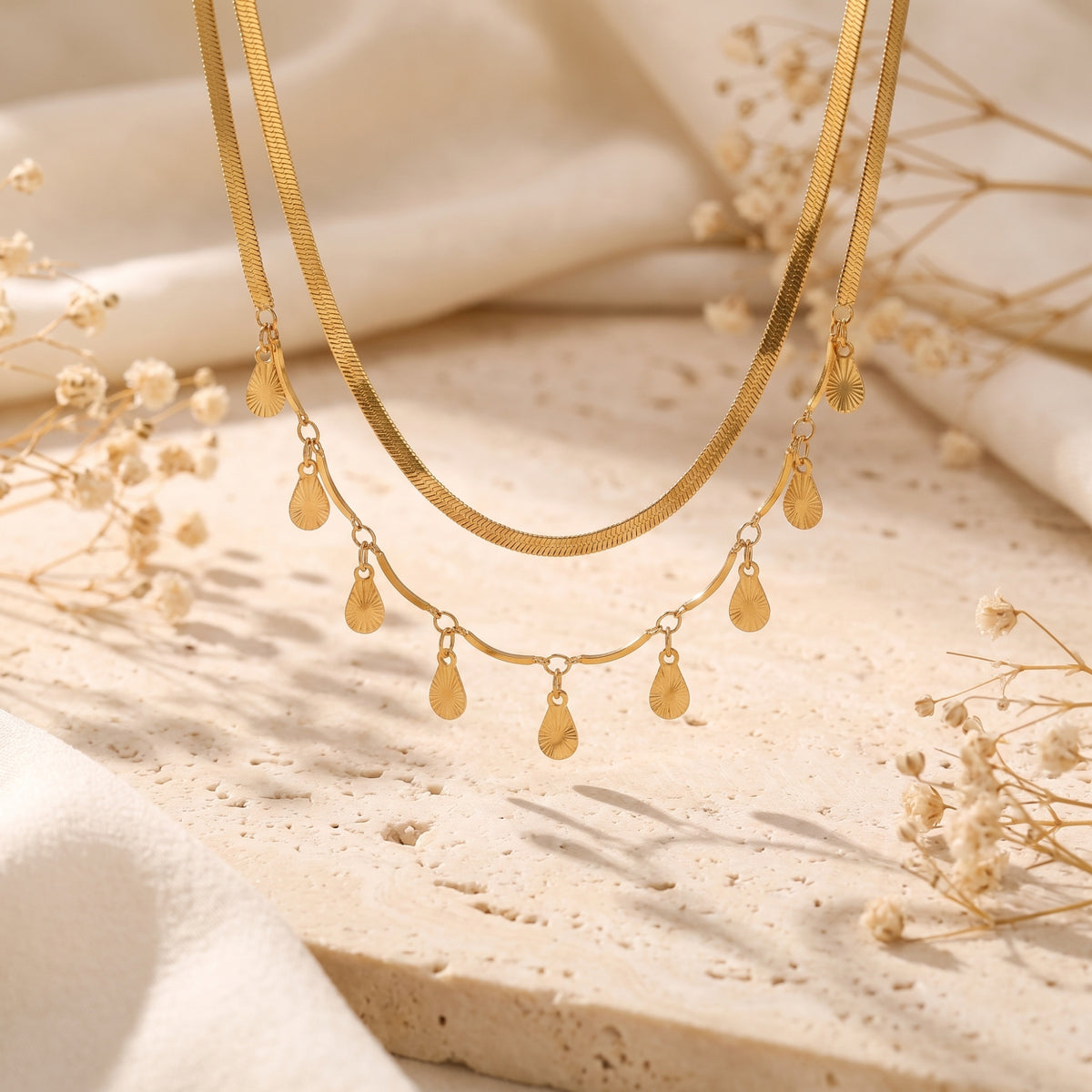 Gold-toned layered necklace hanging vertically, showing a flat snake chain and textured teardrop charms.