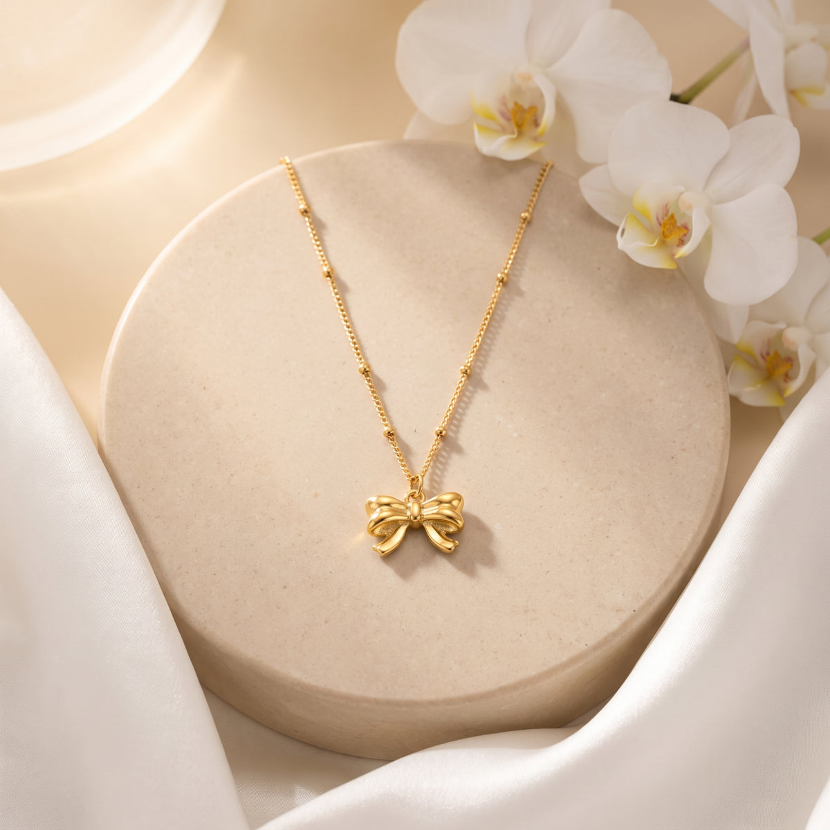 Gold-tone ribbon bow necklace on a beaded satellite chain, displayed on a round beige pedestal with white orchids.