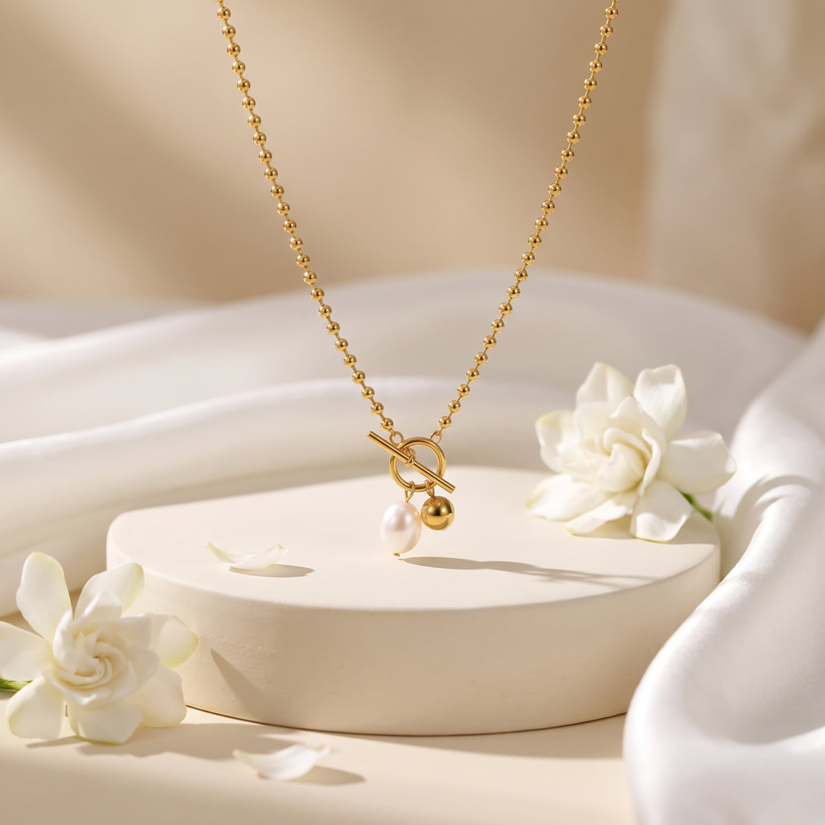 Gold-tone ball chain necklace with front toggle clasp and pearl and sphere charms displayed on a cream pedestal.