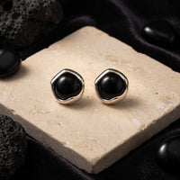 Round black onyx earrings in silver architectural settings on a stone slab with lava rock accents.