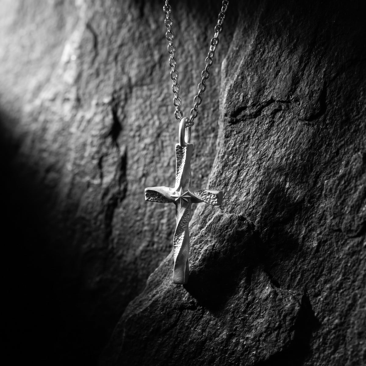 Vertical close-up of a silver twisted cross pendant with starburst engraving hanging against dark textured slate.
