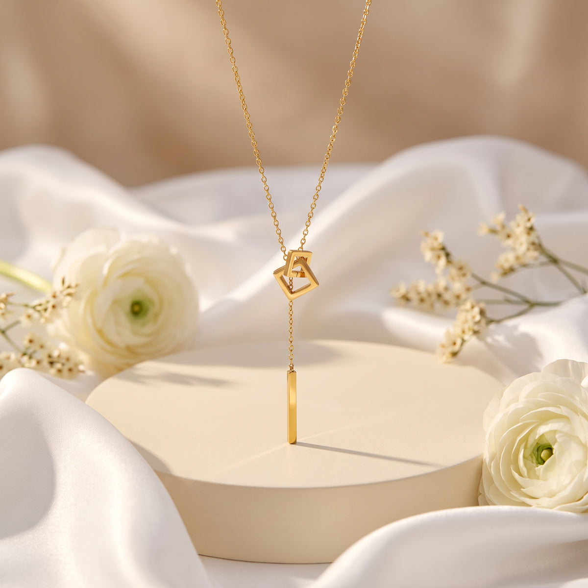 Gold-color square link lariat necklace hanging vertically over a round display stand with white flowers.