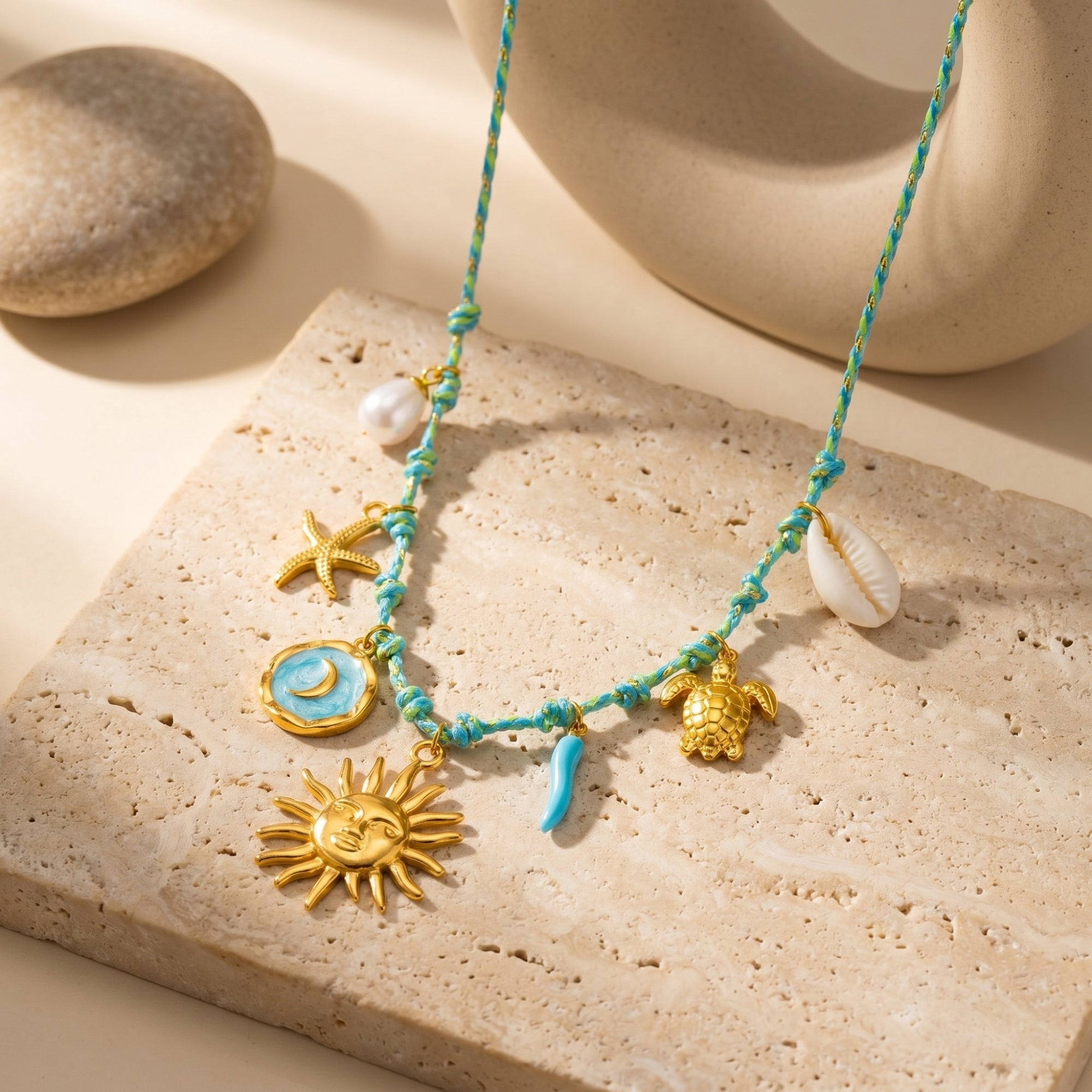 Gold stainless steel charm necklace with pearl and turquoise accents on a blue cord, styled on a stone background.