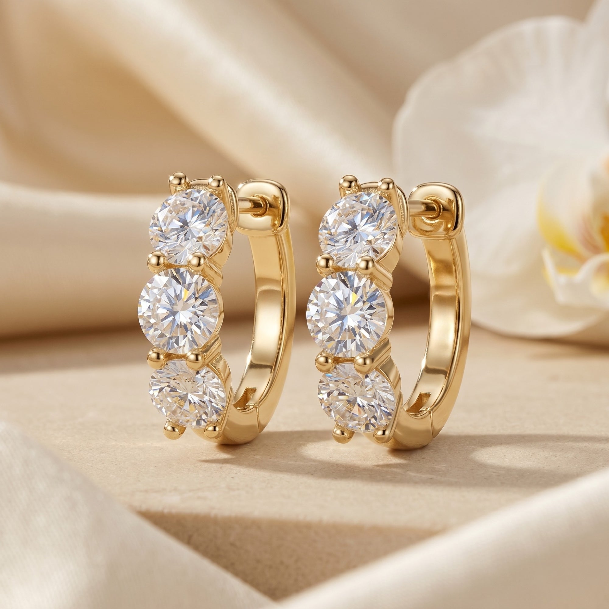 Gold moissanite hoop earrings with three round-cut stones on a soft fabric background.