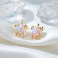 A pair of solid gold turtle stud earrings with oval white opals on white silk fabric.