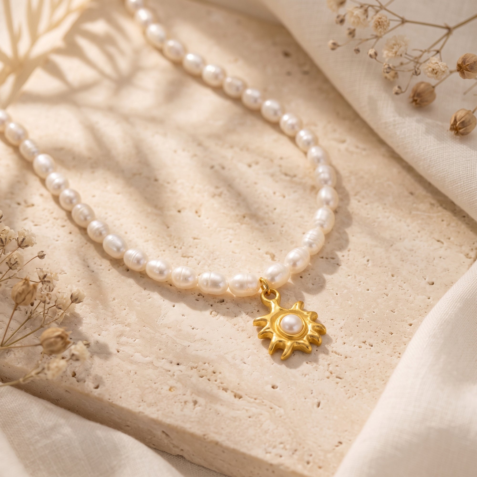 Gold sunburst pendant on a freshwater pearl necklace, styled on a stone surface with dried flowers.