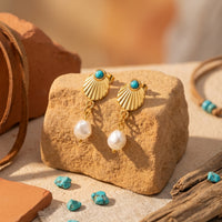 Gold shell drop earrings with turquoise beads and white pearls displayed on a rock in a desert-inspired scene.