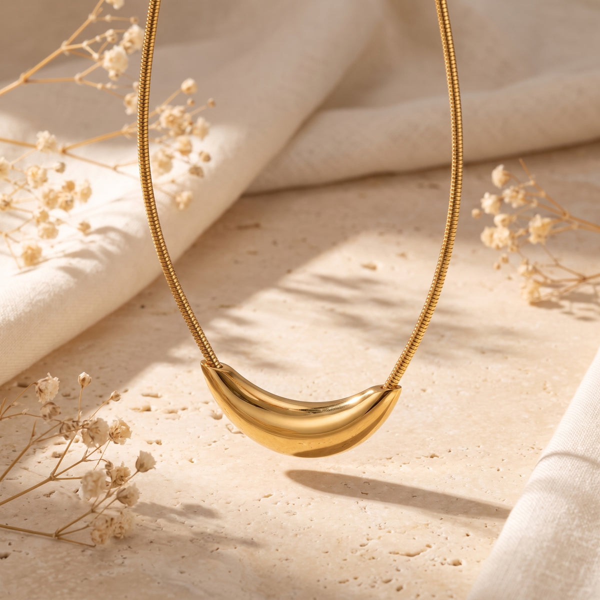 Gold-tone curved crescent pendant on a snake chain suspended over a textured stone surface.