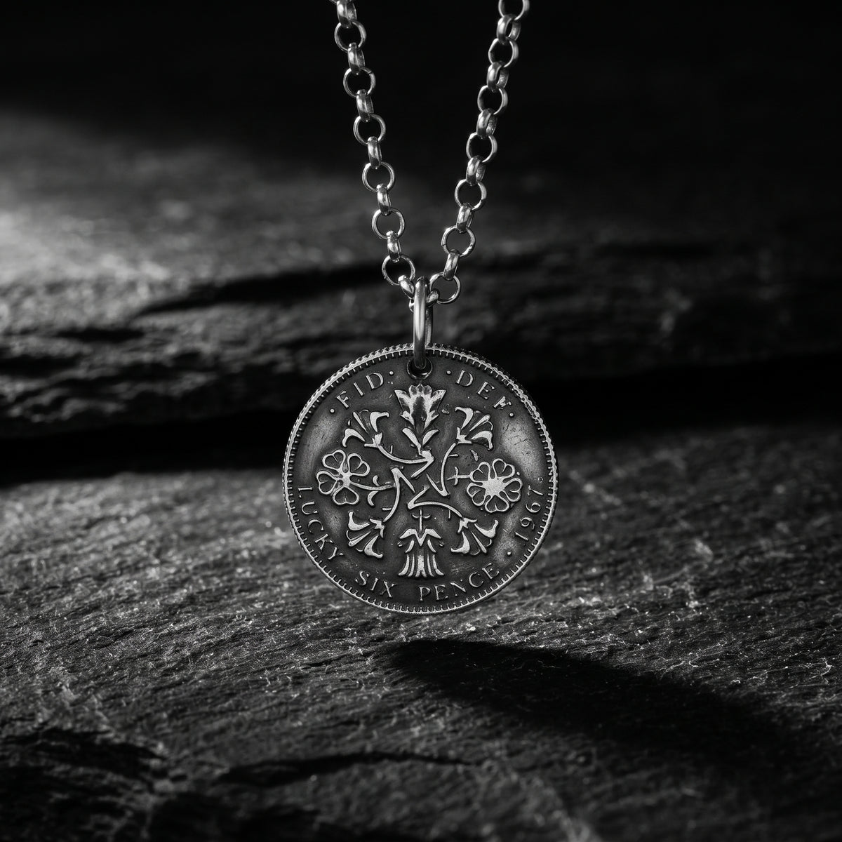 Close-up of a silver coin pendant with floral engravings hanging from a link chain over dark textured rock.