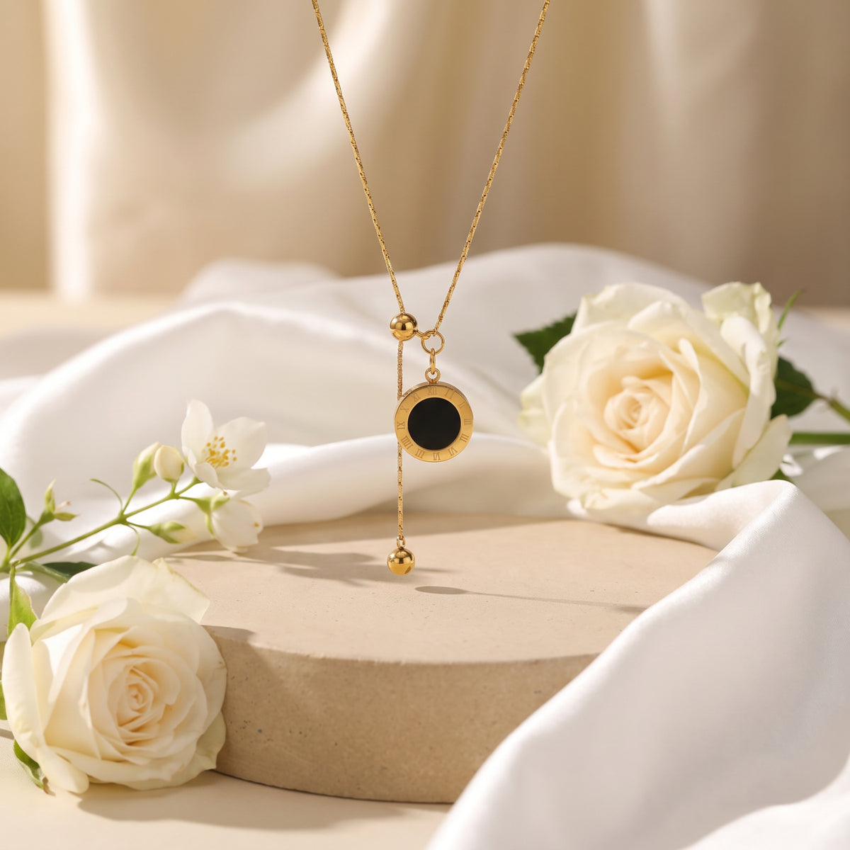 Gold-tone lariat necklace with black Roman numeral disc pendant, displayed over a pedestal with white roses.