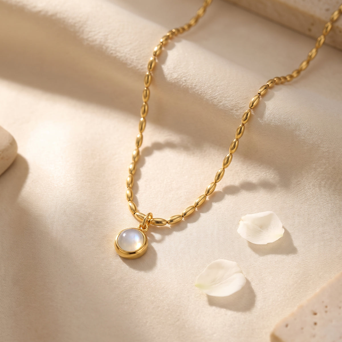 Gold-tone oval-bead necklace with a round bezel-set pendant on cream fabric beside scattered white petals.