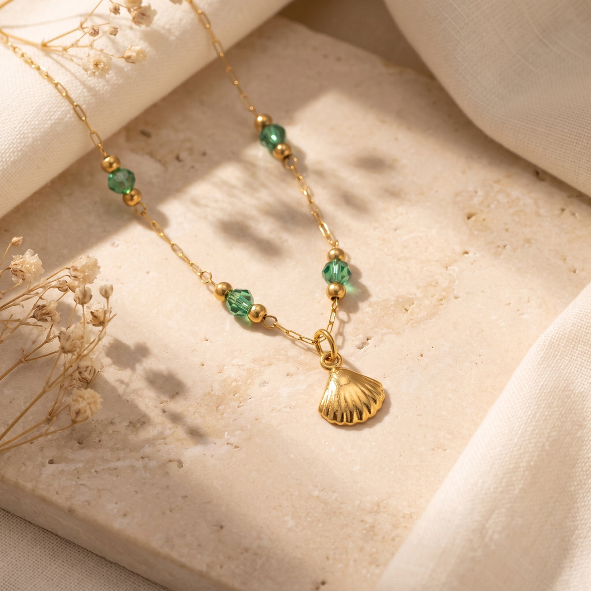 Gold-tone necklace with green crystal beads and a scalloped shell pendant laid on a beige stone surface.