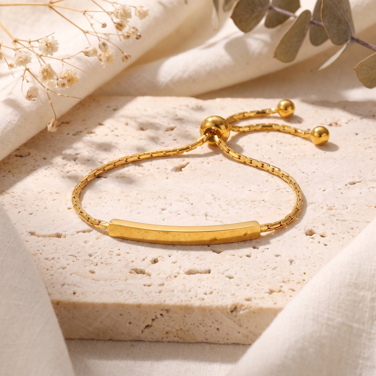 Gold-tone curved bar slider bracelet with a box chain laid flat on a beige stone tile.