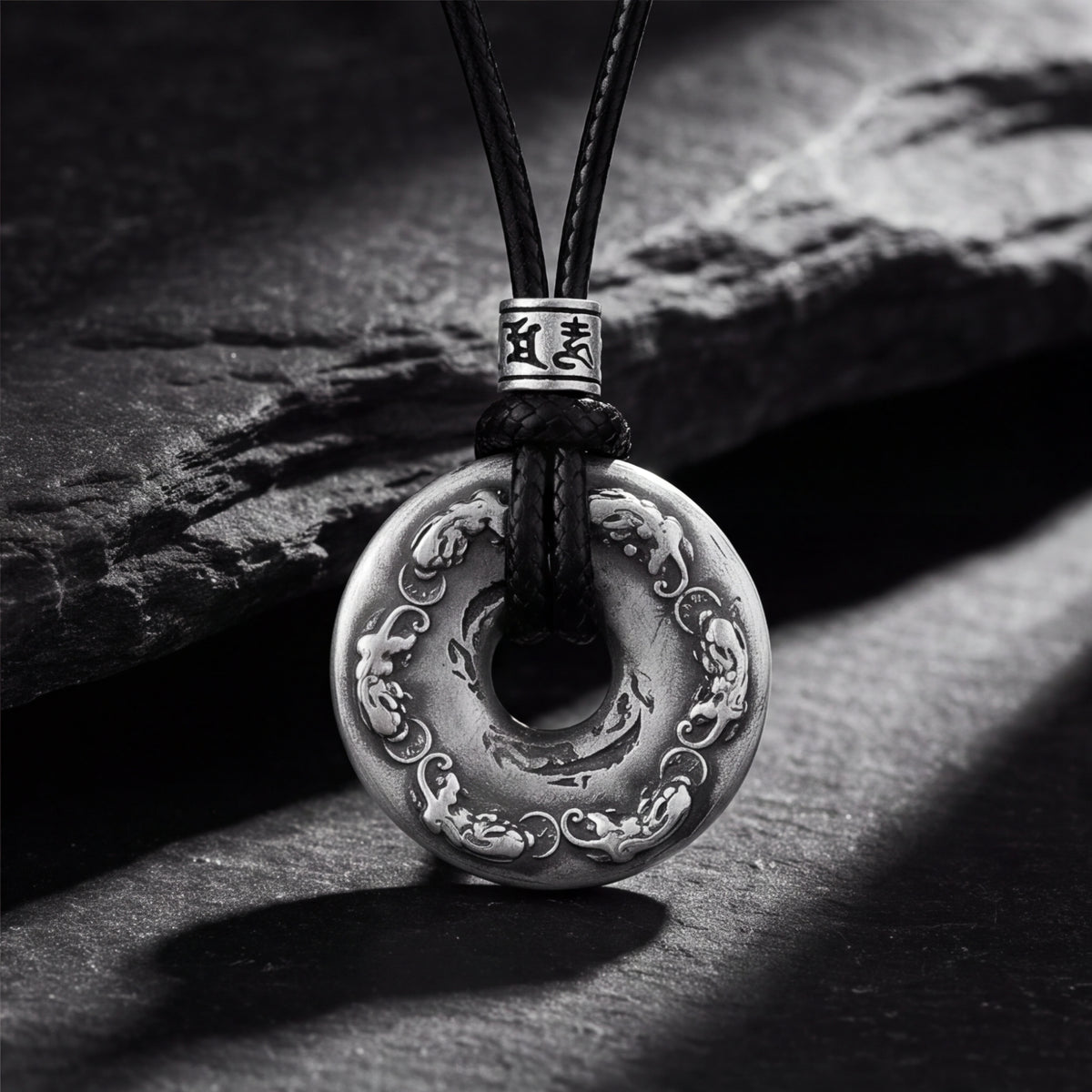 Silver carved disc pendant with relief details hanging from a black cord on a slate background.