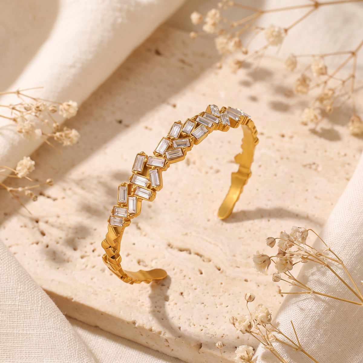 Gold-tone open cuff bracelet with clear baguette-cut stones in a zigzag pattern, standing on a stone slab.