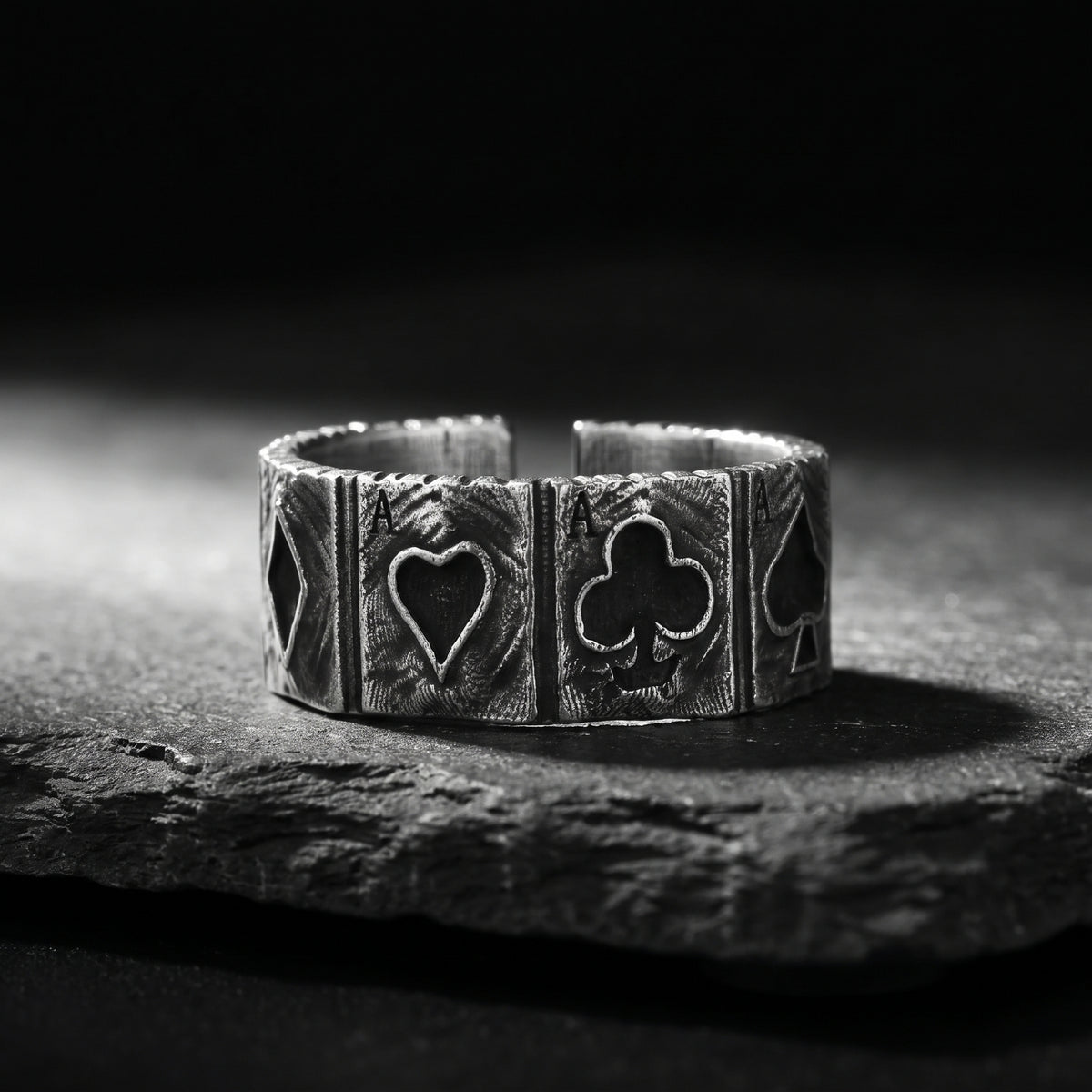 Textured silver open band ring resting on dark slate, showing engraved four-aces playing card panels.